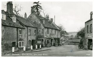 An archive photo labelled, 'Post Office, Bourton on the Hill'. The photo is taken from a road junction and Cotswold stone properties can be seen either side. To the middle left sits the village shop. A sign, 'E I Draper, General Stores' hangs above the door with bay windows displaying produce either side. A low stone wall and views to the hill behind can be seen in the background.