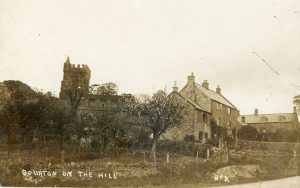 In the centre of the black and white photograph are three linked cottages, later converted into one dwelling, The Stocks (now known as Church House). To the left of the cottages is a garden with small trees. The church of St Lawrence stands in the background. The photograph is labelled 'Bourton on the Hill'