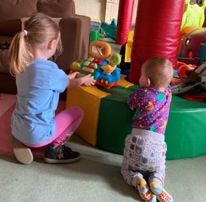 Two young children can be seen from behind. The girls to the left is playing with a brightly coloured plastic cash register toy, The infant next to her has pulled himself up onto a green padded ring and is watching the girl. They are in a playroom with parts of other toys visible.