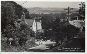 An archive photograph labelled, 'Bourton on the Hill, The Cotswolds from the Hill Top'. It shows the main road through the village with views looking out over Moreton in Marsh and the countryside beyond. A 'Drive Slowly' sign is positioned on the left. Mature trees obscure most of the buildings on either side of the road. Telecommunication poles and cables are visible, flanking the road.