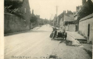 An undated historic photo, circa 1920, looking up the main road of Bourton on the Hill with overhead telecommunication cables positioned along it. There is an old fashioned motorbike and sidecar, containing a passenger, parked next to a still present water trough. The tower of St Lawrence's Church can be seen in the distance.