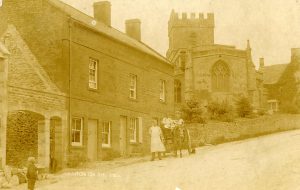 An archive photograph 'labelled Bourton on the Hill'. St Lawrence Church can be seen behind a wall lining the road. A man in a long white apron is standing in front of the houses to the left of the church. Next to him is a horse and cart with gentlemen sitting atop. Their clothing suggests the picture was taken in the early 1900s. They appear to be on their delivery round. A small boy is visible, standing in the road, facing away from the camera.