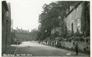 An undated historic photo, circa 1920, looking up the main road of Bourton on the Hill towards the Horse and Groom pub with the school to the left and cottages to the right. A number of children line both sides of the street.
