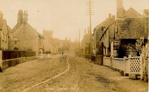 An old photograph looking up the main road in Bourton on the Hill. The road is unsurfaced and wheel marks are visible within it. Stone cottages and houses are on either side of the road and telecommunication poles are positioned along the right hand side.