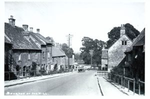 An undated historic photo looking down the main road in Bourton on the Hill, with overhead telecommunication poles and lines. An early motor truck is visible on the left side of the road.