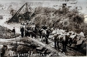 Historical black and white photo. Three horses pulling a wooden cart of stone from a quarry. Two men are alongside them