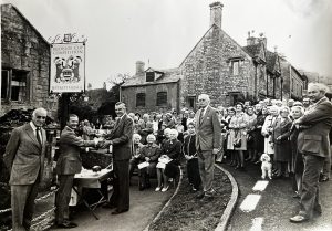 A black and white image showing the presentation of a cup. Many people are sitting and standing behind, watching the proceedings. An impressive sign reads - Bledisloe Cup Competition, Best Kept Village. 1977